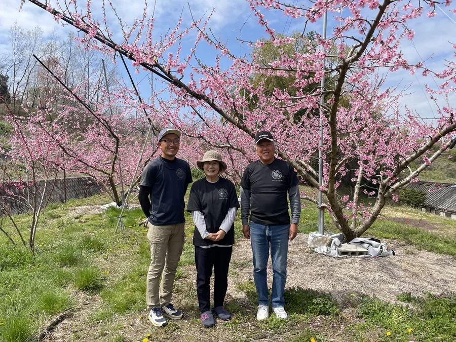 桃の花が咲く園地に立つ萬亀雅農園の皆さん